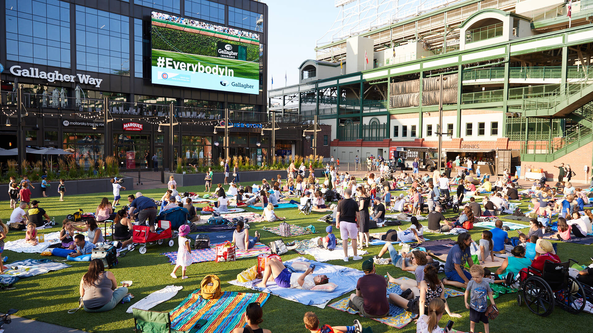 Families and friends on blankets watching Movie Night on the Gallagher Way lawn with the Wrigley Field video board visible